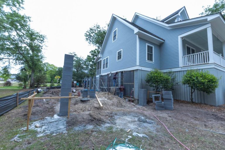 Concrete block column going up beside a raised blue house with a covered porch, with block stacks and string lines on the site