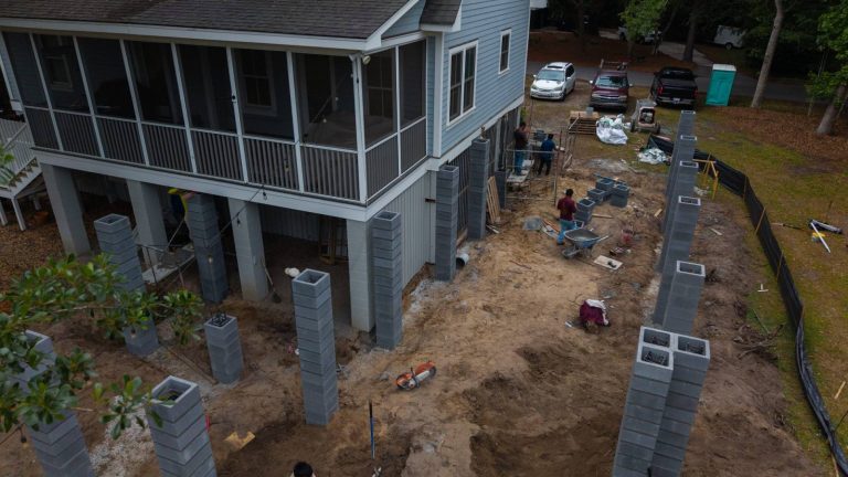Raised blue home with a screened porch beside rows of new hollow block columns for an addition, seen from above